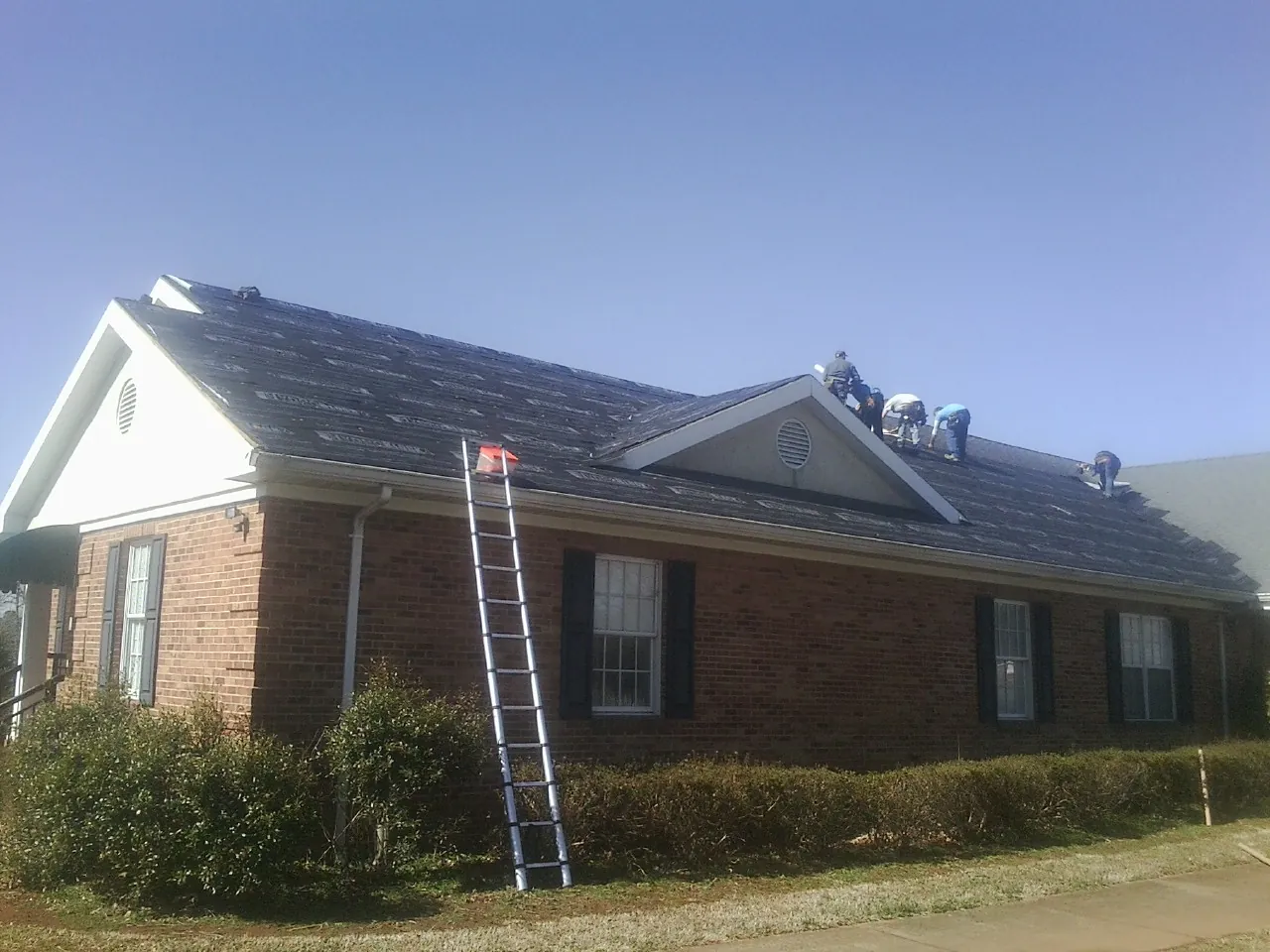 Crew tearing shingles off a brick ranch roof with a ladder set — Metal Roof Repair work by our crew in Flintville
