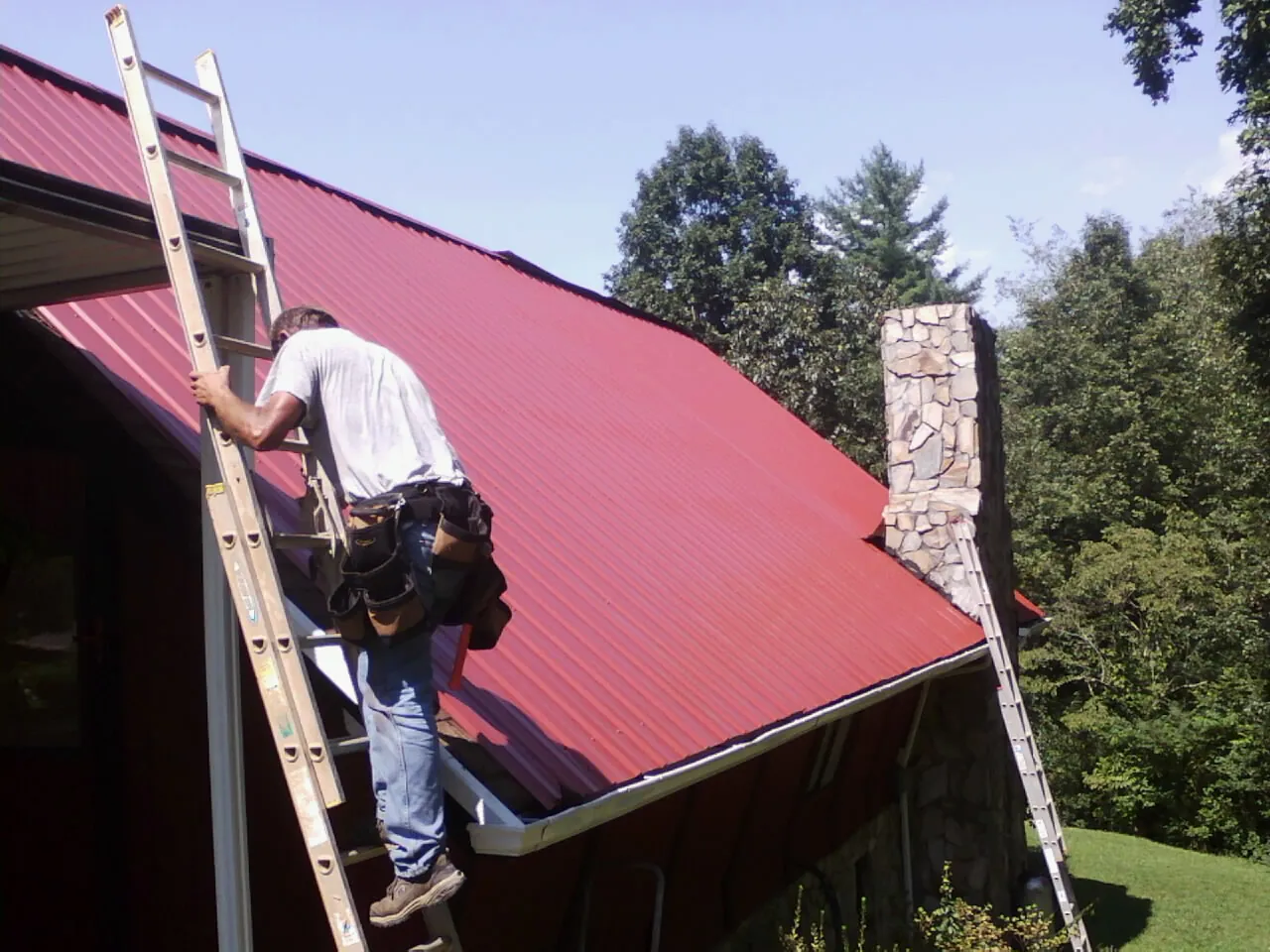 Roofer on a ladder beside a red metal roof and stone chimney — Soffit & Fascia Repair by Roofing Brothers in Flintville